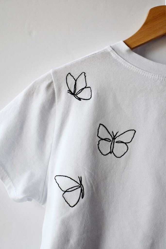 हाथ से कढ़ाई की गई तितलियों वाली सफ़ेद टी-शर्ट। काली कढ़ाई वाला धागा, केवल रूपरेखा