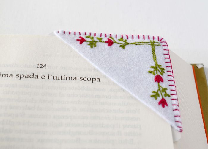 हाथ से कढ़ाई की गई कोने वाली बुकमार्क जिसमें किताब पर छोटे-छोटे फूल रखे गए हैं